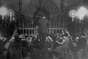 Schwarz-weiß-Foto eines Orchesters mit Dirigent in einem großen Saal bei einem Konzert