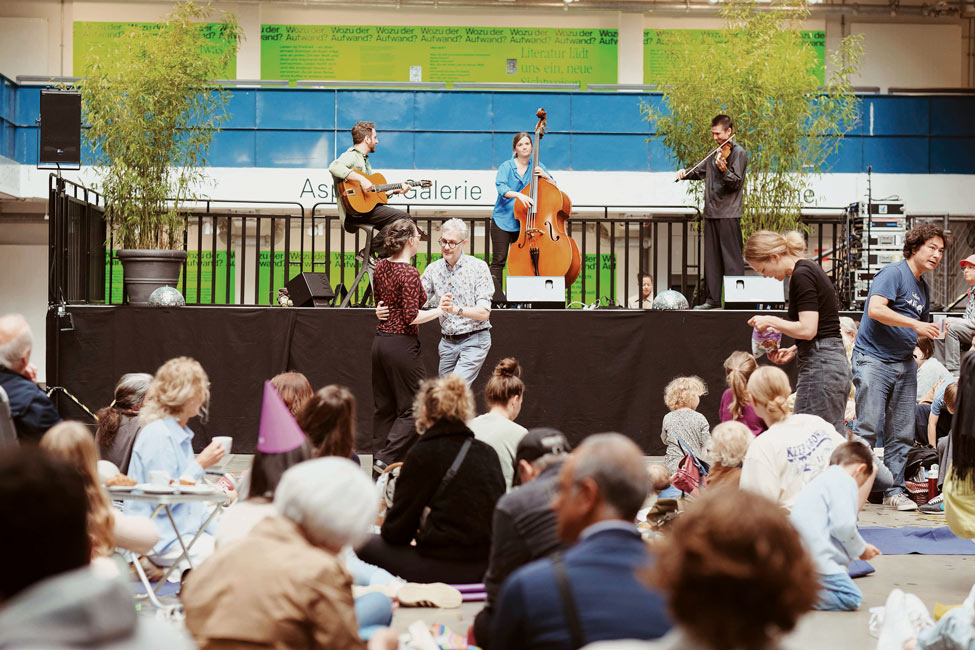 Drei Musiker*innen spielen Gitarre, Kontrabass und Geige auf einer Bühne vor sitzendem und tanzendem Publikum in einer hellen Halle.