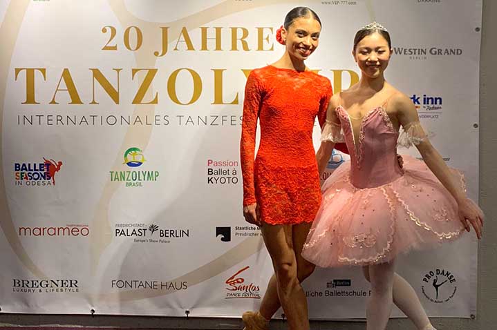 Die Tänzerinnen Rachel Pimentel Quintao Maika Kumazaki stehen vor einem Werbebanner des Tanzfestivals Tanzolymp