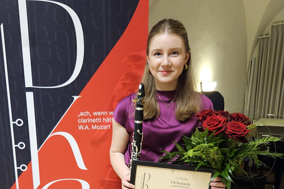 Porträt von Veronika Troiber vor dem Banner der Deutschen Klarinetten-Gesellschaft. Sie hält ihre Klarinette, einen Blumenstrauß und die Urkunde in den Händen.