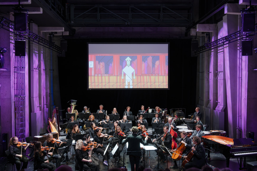 Orchester mit Dirigent vor einer Leinwand in einer Halle mit violett beleuchteten Säulen