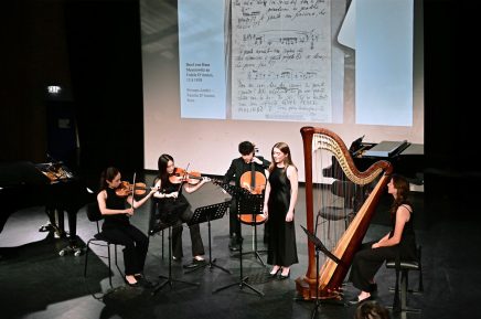 Fünf Musikerinnen und Musiker auf einer Bühne mit Streichinstrumenten und Harfe vor einer Leinwand mit handschriftlichen Noten und Notizen.