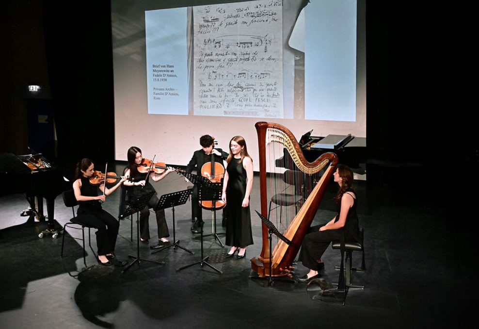 Fünf Musikerinnen und Musiker auf einer Bühne mit Streichinstrumenten und Harfe vor einer Leinwand mit handschriftlichen Noten und Notizen.
