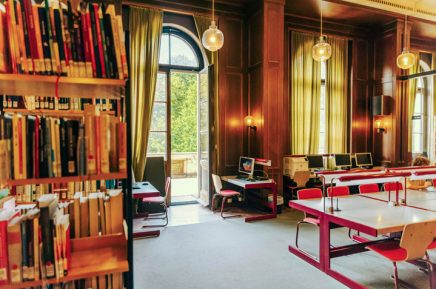 Großer Lesesaal einer Bibliothek mit hohen Fenstern, langen Vorhängen, Bücherregalen und Tischen mit roten Stühlen.