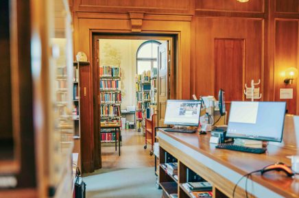 Innenraum einer Bibliothek mit Holzregalen, Computern auf einem langen Tisch und Blick durch eine Tür zu weiteren Bücherregalen.