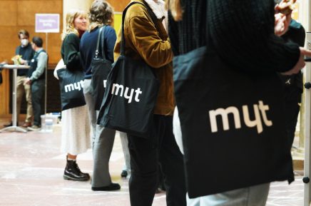 Mehrere Personen stehen in einer Reihe und tragen schwarze Stofftaschen mit weißer Aufschrift 'myt'