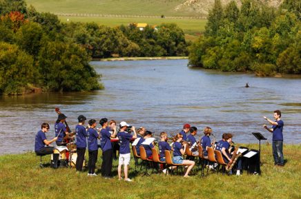 Blasorchester spielt im Freien am Flussufer mit Dirigent und Musikern in blauen T-Shirts