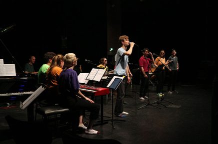 Gruppe Studierender auf der Bühne an Instrumenten und ein Sänger am Mikrofon im Mittelpunkt