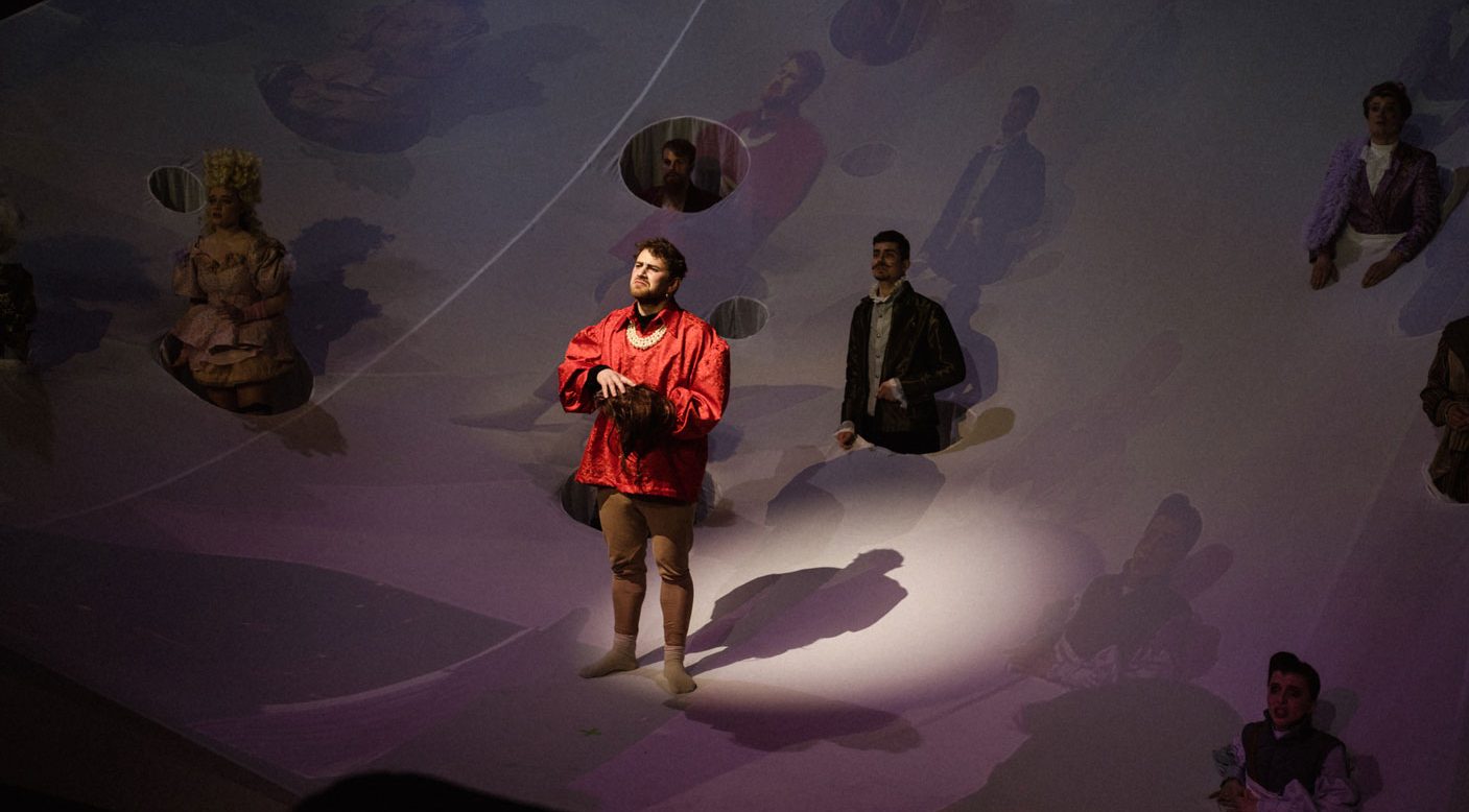 Ein Mann in barockem Kostüm steht auf einer Bühne, die mit einem Tuch überdeckt ist.